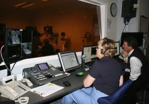 Two people in a control room looking through window at a simulation training.