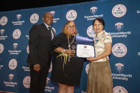 Photo of three individuals standing on stage at a ceremony. On the left is a man in a suit, in the middle is a woman holding an award and a certificate, and on the right is a woman in a traditional outfit holding a certificate. The background is a banner that says ‘Samuel Merritt University Health Impact Awards.'