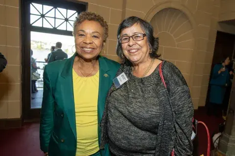 Photo of two women smiling for a photo. The woman on the left is wearing a green blazer with a yellow shirt, and the woman on the right is wearing a black top with a gold necklace. They are posing in front of a window with a view of the outdoors.