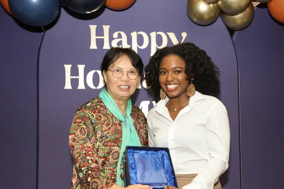 President Wang and student, Victoria Campfield hold an award in front of a blue happy holidays sign with balloons.